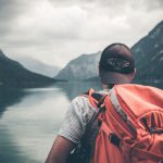 Travel - man with red hiking backpack facing body of water and mountains at daytime